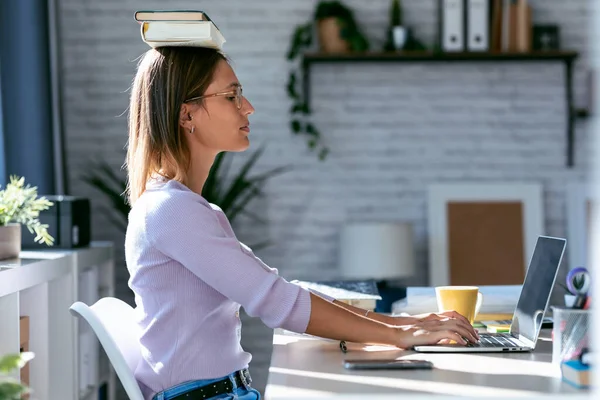 Shot of young attractive woman at the desk with books on her head while working with computer at home.