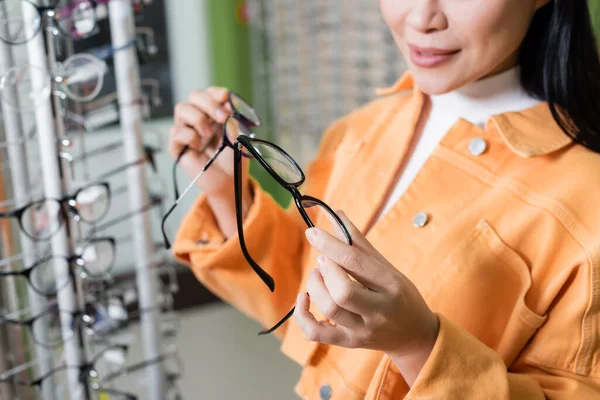 cropped view of blurred woman comparing eyeglasses in optics shop